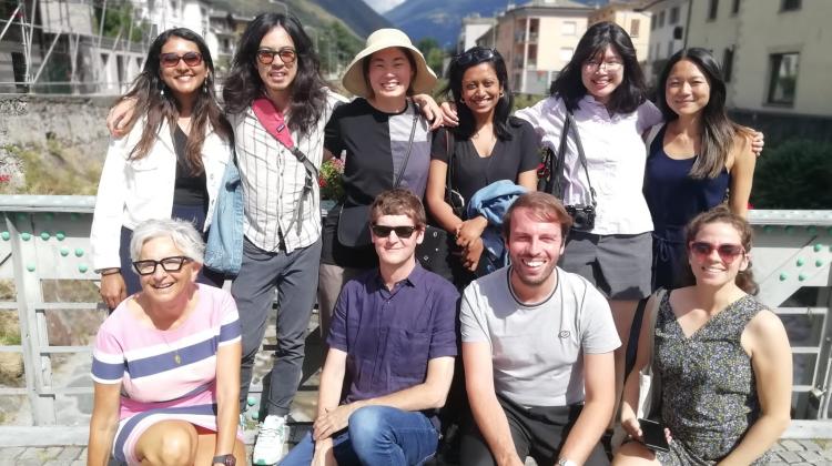 Group photo of students and instructors. Behind the group one can see a sprawling Italian town, laid out descending a mountain. In the far background, the mountain ranges can be seen rising above the group. 