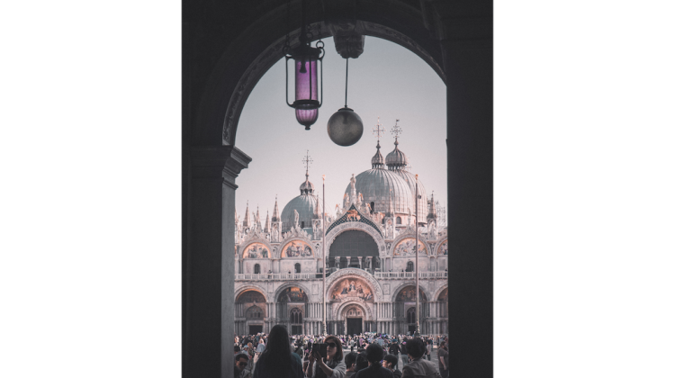 A group of tourists snaps some selfies from the ground floor of the former Palazzo Reale at the west end of Piazza San Marco, looking out from the deep shade of the classical arcade.